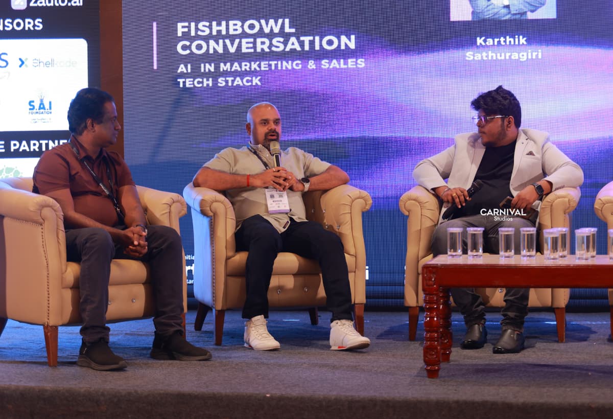 Maran on a panel of three in armchairs, holding a microphone, under a screen reading “Fishbowl Conversation”